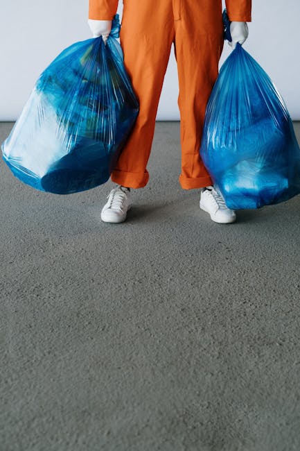 A person wearing orange overalls and white sneakers is standing on a grey carpeted floor inside a property, holding two large blue plastic bags filled with household waste or bulky items used for home