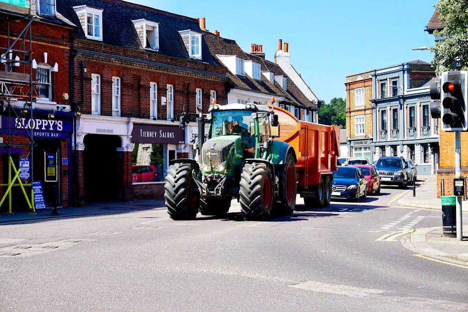 A tractor with an orange and green trailer is positioned at an intersection on Ruislip High Street during daytime, with a clear blue sky overhead. The tractor is parked next to a row of brick and Vict